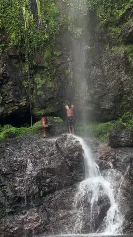 A person is standing under a waterfall. They are wearing dark clothing. The waterfall cascades down a rocky surface into a small pool. Behind the person is a rocky cliff covered in greenery. To the left of the person is a small orange and black object.