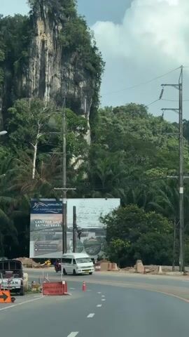The video shows a roadside view with a large, sheer rock face covered in lush greenery in the background. In the foreground, there is a paved road with traffic cones and barriers indicating construction or roadwork. Several vehicles are visible, including a white van in the center lane, a black SUV to the left, and a white car further down the road. There are also utility poles with electrical wires running across the frame. Two large billboards are prominently displayed on the left side of the road, one advertising "FLOURISH VILLAS KRA" with details about land for sale, and another with Thai text and images. The sky is partly cloudy. The overall scene depicts a developing area with infrastructure, commercial advertising, and a striking natural landscape.
