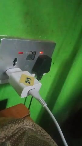 The video shows a white power adapter plugged into a wall outlet with a green wall behind it. A white USB cable is connected to the power adapter. The outlet has red indicator lights illuminated. Next to the adapter, a black plug is inserted into another outlet. A brown patterned surface is visible below the outlet. A yellow sticker with the number "219" is affixed to the power adapter.