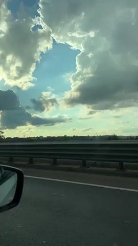A video shot from inside a car, looking out the passenger window. The camera pans slightly, capturing a highway scene. In the foreground, the passenger-side mirror is visible, reflecting a white car and a bit of the sky. The road surface is asphalt with a white line dividing lanes. A metal guardrail runs along the side of the road, separating it from a body of water or land beyond. In the distance, a tall, slender pole stands against a sky filled with dramatic, fluffy white and grey clouds. Sunlight breaks through the clouds in some areas, creating a bright, diffused light. The overall impression is a view from a moving vehicle on a cloudy day.