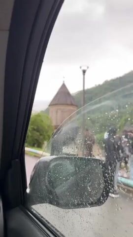 The video, shot from inside a car, shows a view through a wet car window of a stone building with a conical roof and a cross on top. The building is surrounded by lush green trees. In the foreground, a side mirror of the car is visible, also covered in water droplets. The sky is overcast and appears to be raining, as indicated by the water on the window and the presence of umbrellas held by figures blurred in the background. A streetlamp can be seen on the right side of the frame, and a hillside is visible in the distance. The overall atmosphere is subdued due to the rainy weather.