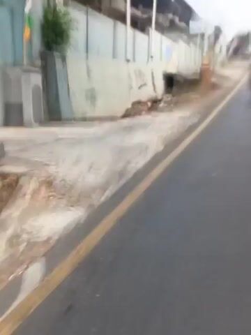 The video shows a first-person perspective of a motorcycle moving along a paved road with a yellow line marking the edge. The road is bordered by a dirt and gravel area to the left, which then leads to a low concrete wall covered in graffiti. Beyond the wall are buildings, some appearing to be shops or houses with light-colored facades and some darker structures. A golden-topped monument is visible on the left side of the road. The environment appears to be an urban or semi-urban setting, possibly in a developing country, with some debris and uneven terrain visible. The video is short, only lasting a few seconds, and the motion of the motorcycle creates a blurred effect.
