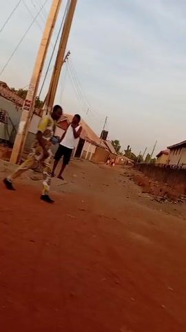 The video shows a street scene in a residential area. The street is unpaved and has a reddish-brown color. Power lines and utility poles are visible overhead. A man in a white tank top and dark shorts is walking in the street. Another person, wearing a yellow and patterned outfit, is walking in the opposite direction. The camera pans to show a wall with barbed wire on top, a closed metal gate, and a building with barred windows. The scene appears to be in a developing country. The sky is overcast.