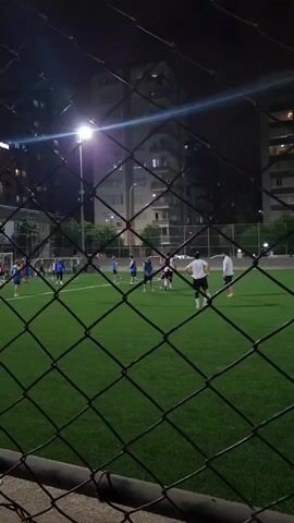 A group of people are playing soccer on a bright green artificial turf field at night. They are illuminated by stadium lights. A chain-link fence is in the foreground, obscuring part of the view. Two buildings are visible in the background.