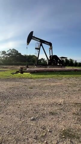 The video shows an oil pumpjack operating in a field under a clear blue sky. The pumpjack is black and has a large counterweight. It is positioned on a concrete pad surrounded by grass and dirt. There are some pipes and equipment near the pumpjack. The sun is setting or rising, casting a warm light on the scene. The video is shot from a low angle, looking up at the pumpjack. The pumpjack is in motion, with its beam moving up and down. The sound of the pumpjack is audible.