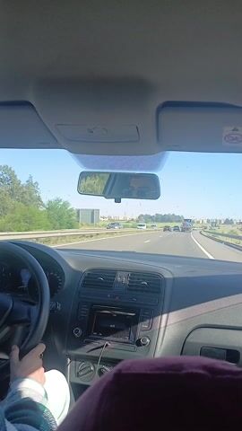 Here is a description of the video.

The video shows a first-person perspective from inside a car driving on a highway. The sky is clear and blue. The driver has their hands on the steering wheel. Several other vehicles are visible on the highway, including a truck. The dashboard of the car is visible, including the radio. The back of another person's head is visible in the foreground. There are trees and buildings visible in the distance, adjacent to the highway.