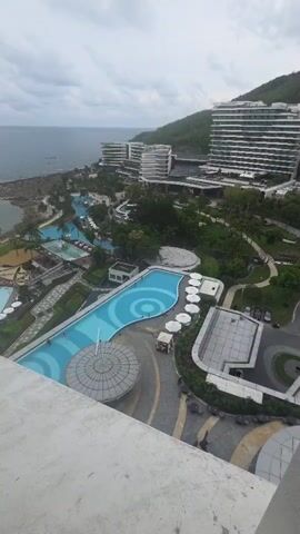 This aerial video shows a resort complex on a cloudy day. The main subjects are the numerous buildings, swimming pools, and the surrounding landscape. The resort features several multi-story white buildings with modern architecture, a large winding swimming pool with blue and white circular patterns, smaller pools, palm trees, and lush greenery. In the distance, the ocean meets a rocky shoreline, and a hill covered in vegetation rises behind the resort.

The primary activity observed is people enjoying the resort's amenities. Several individuals are visible swimming and relaxing in the main pool. There are also lounge chairs and umbrellas set up around the pools, suggesting leisure activities. The scene depicts a typical day at a holiday resort, focused on relaxation and recreation.

Notable details include the overcast weather, indicated by the cloudy sky, which suggests it might be morning or late afternoon. The location is identified as Jiyang District, China. The overall mood is peaceful and serene, characteristic of a vacation destination. No visible text is present in the video. The video provides a sweeping view of the resort, highlighting its scale and design.