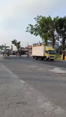 The video pans across a street scene. On the left side of the road, there is a sidewalk made of interlocking hexagonal pavers, followed by a paved road. Several motorcycles are visible on the road, some of which appear to be moving in the same direction as the camera's pan. On the right side of the road, there is a large yellow truck with a white cargo container. Beyond the road, there are buildings, trees, and utility poles. The sky is overcast. There are several people visible on motorcycles.