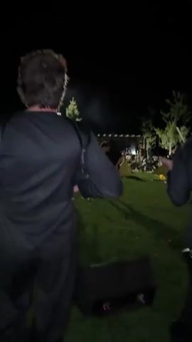 The video shows two men in a grassy outdoor area at night. The first man, seen from behind, is wearing a black jumpsuit and appears to be carrying some equipment on his back. He turns towards the camera, revealing his face and a beard. He is holding a violin and bow and begins to play. In the background, another man in dark clothing is walking away from the camera. The scene is dimly lit, with some artificial lights illuminating the trees and structures in the background. The first man then puts down the violin and picks up an accordion, which he starts to play.