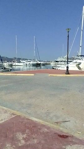 The video displays a marina with several sailboats docked under a clear blue sky. The camera is positioned at a low angle, looking upwards. The foreground shows a paved walkway with some green and purple splotches, possibly from paint or chalk. A lamppost stands near the edge of the walkway, overlooking the water. The boats, predominantly white with tall masts, are lined up in the background. The overall impression is a sunny day at a nautical location.