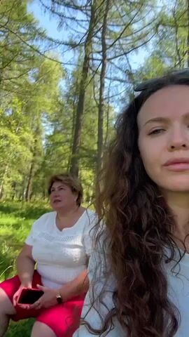 Two women are in a park setting. The woman in the foreground, on the right, has long, curly brown hair and is speaking into the camera, appearing to be recording a video. The woman in the background, on the left, has short, light brown hair, is wearing a white top and red shorts, and is sitting down. She is looking to her left and holding a red smartphone. The background consists of tall trees and green foliage, suggesting a sunny day. The woman in the foreground asks the woman in the background if she likes it there, and the woman in the background responds positively.