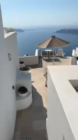 The video shows a panoramic view of a coastal landscape in Santorini, Greece. The foreground features white-washed buildings with smooth curves, typical of the island's architecture.  Outdoor seating areas with tables, chairs, and umbrellas are arranged on terraces.  The deep blue Aegean Sea stretches out to the horizon, with a volcanic caldera and other islands visible in the distance. The sky is clear and blue, indicating a sunny day.