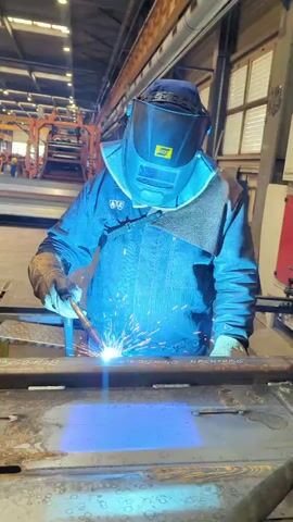 A person in a blue welding jacket, protective helmet, and gloves is welding a metal beam. Sparks are flying from the welding torch. The welding area is illuminated with blue and white light. The background appears to be a factory or workshop with large windows and orange industrial equipment.