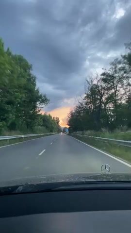 This is a first-person perspective video of a car driving on a road at dusk. The sky is cloudy with hints of orange and pink from the setting sun visible in the distance. Trees line both sides of the road, and a guardrail is visible on the left. The car's dashboard, including a Mercedes-Benz logo, is visible in the foreground. The road is paved and has white lane markings. There are other vehicles visible in the distance on the road.