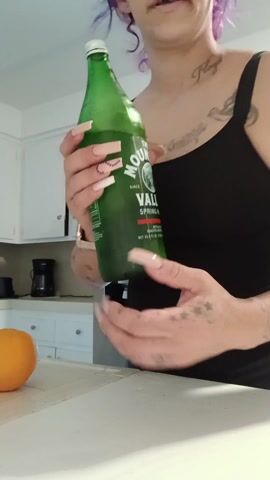 A woman stands in a kitchen and holds a green glass bottle of The Mountain Valley Spring Water. She wears a black tank top and gray pants. Her hair is a purplish color, and she has tattoos on her chest and arms. On a white counter in front of her sits an orange. Behind her are white kitchen cabinets and a stainless steel refrigerator. She struggles to open the bottle with her fingernails.