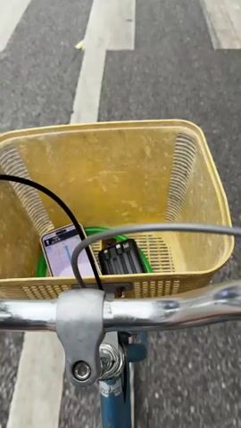 The video begins with a view inside a bicycle basket, which contains a smartphone displaying a map, a black battery pack, and a green lock. The camera then shifts to a first-person perspective of someone riding the bicycle down a wide, gray asphalt road. Buildings line the street on both sides, with shops and restaurants visible. Mountains appear in the distance. Various vehicles, including cars, motorcycles, and scooters, move along the road. The road is marked with white lines, and some areas of patching and repair are evident. People are walking and riding scooters along the sidewalks. The sky is overcast.