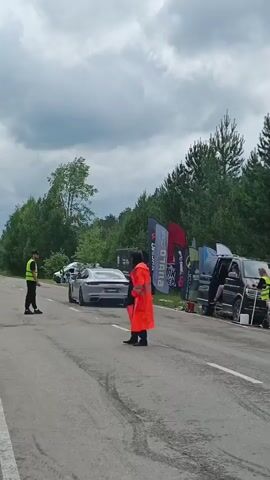 A car race is taking place on a paved road. Two people are standing on the road, one in a neon yellow vest and the other in an orange rain coat. The person in the yellow vest is standing on the left side of the road, and the person in the orange rain coat is standing in the middle of the road. Both appear to be directing traffic or signaling. A white sports car is visible in the distance. There are several tents and flags set up on the right side of the road, indicating a spectator or event area. The environment appears to be a rural or wooded area, with trees visible in the background. The sky is cloudy.