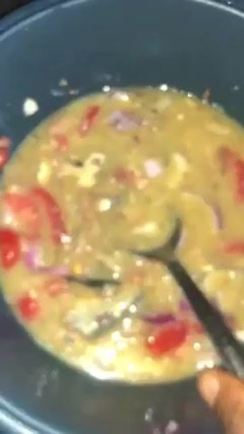A close-up, slightly blurred video shows a dark pan containing a light brown liquid with visible red and purple pieces. A metal spoon stirs the mixture continuously. The pan appears to be sitting on a flat surface.