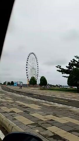 The video shows a large Ferris wheel in the distance. The camera is inside a vehicle moving along a road or walkway. A man is walking in the same direction as the vehicle, pushing a stroller. There are trees and poles lining the road, and a "no parking" sign is visible. The sky is overcast.