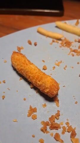 The video shows a close-up view of a blue plate with remnants of food. A single fried food item, resembling a mozzarella stick or a chicken tender, is positioned in the center of the plate. The item is golden brown and appears to be breaded. Surrounding the food item are scattered crumbs, likely from the breading. Two elongated, pale yellow food items, possibly breadsticks or fries, are visible in the upper portion of the plate. To the right of the main food item, a piece of white, possibly starchy food, is also present. The plate is resting on a wooden surface, suggesting a table. The background is slightly blurred but indicates an indoor setting. A voice is heard speaking in Indonesian, but the content is not clearly discernible. The overall scene depicts the aftermath of a meal.