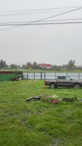The video opens with a wide shot of a yard on a gray, overcast day. In the foreground is a patch of wet grass with some yellow flowers. Behind that, a darker green fence encloses a yard with a pickup truck parked on the right. Across the street, there are houses with red roofs and a few trees. The video then pans to the left, revealing more of the yard and houses. A wooden porch railing with a mug on it is visible on the right side of the frame. The sky is a uniform gray, and the overall impression is one of a rainy or misty day in a rural setting. There are no people visible in the video.