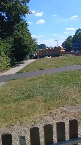A tractor pulls a trailer loaded with hay bales along a road. The tractor is partially visible on the left side of the frame, pulling the trailer which is filled with numerous round hay bales. The trailer is moving towards the right side of the frame. The road appears to be in a rural or semi-rural area, with trees and grassy areas lining the sides. The sky is blue with scattered clouds, indicating daylight. There are a few cars visible in the distance on the right side of the road, suggesting it is a public road. A blue road sign with a white symbol is also visible in the distance on the right. The foreground shows a patch of grass and a paved pathway.