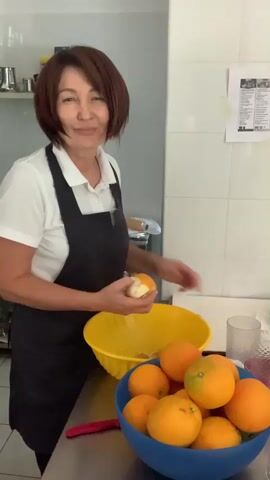 A middle-aged woman with shoulder-length brown hair, wearing a white polo shirt and a black apron, stands in a kitchen. She is smiling and winking at the camera while peeling an orange. In front of her, on a stainless steel counter, are two bowls filled with oranges. One bowl is yellow and the other is blue. There is also a red object that appears to be a knife or a peeler to the left of the yellow bowl. The background shows white tiled walls, some shelves with kitchen equipment, and a notice pinned to the wall.