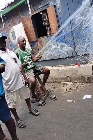 The video shows three men in an outdoor setting. A man sits on a wooden stool mending a large fishing net. He is wearing a green Boston Celtics jersey and shorts, and sandals. He occasionally looks toward the camera with a smile. Standing beside him is a man wearing a white shirt, khaki shorts, and a baseball cap. To the left, another man stands wearing a dark shirt and blue shorts.  The background features a blue and green painted building with a small closed window. The area around them is paved and there are pieces of discarded rope and small trash on the ground.