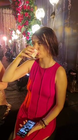A woman in a pink dress is drinking from a paper cup while holding a smartphone in her left hand. She is standing near a decorative backdrop of flowers and lights. There are other people in the background and what appears to be event staging equipment. The scene has a celebratory or festive atmosphere.
