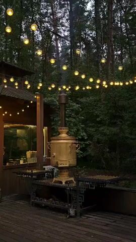 The video captures an outdoor scene at dusk, likely in a forested area, illuminated by string lights. In the center, a large, ornate brass samovar is prominently displayed on a decorative metal stand. Smoke gently rises from the samovar's chimney, suggesting it's in use. To the left, a wooden structure, possibly a building or covered patio, is visible, with more string lights extending from it. A wooden deck forms the foreground. A young child, wearing a jacket and jeans, walks across the deck from the left side of the frame towards the center, pausing briefly. The background consists of a dense wall of trees, their dark forms creating a natural backdrop. The overall atmosphere is cozy and inviting, with the warm glow of the lights contrasting with the dim natural light of the evening.