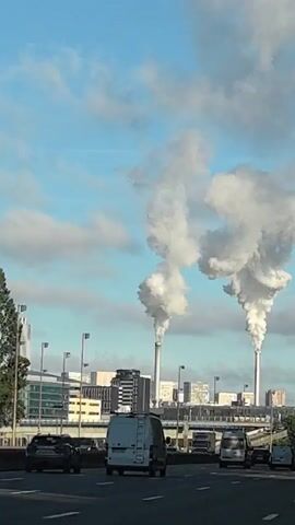 The video shows a view from a highway looking towards a city skyline. Two tall smokestacks are visible in the distance, emitting large plumes of white smoke. The sky is blue with scattered clouds. Several cars are driving on the highway in the foreground, including a white van. Buildings and infrastructure, such as overpasses and streetlights, are visible in the city. The overall scene suggests an industrial area within or near an urban environment.