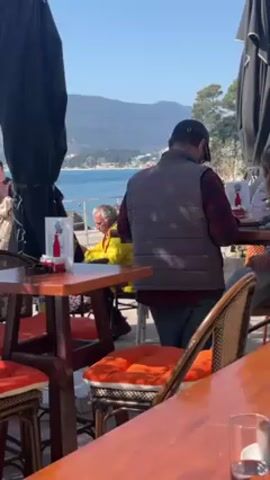 The video captures a sunny day at a seaside cafe, where several people are seated at tables. The cafe appears to be located along a promenade, as the sea and distant hills are visible in the background. The tables are made of wood, and the chairs are woven, with orange cushions on the seats.  One man is standing, and another man is seated at one of the tables. Other patrons are walking in the background.  Large black umbrellas provide shade to some tables. The overall atmosphere is relaxed and leisurely.