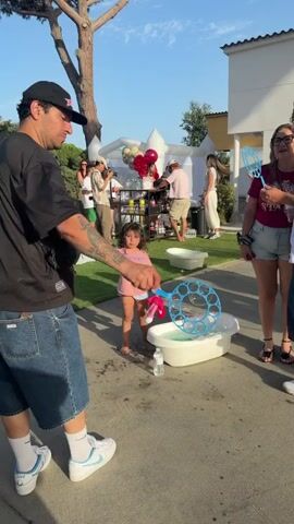 A man wearing a black t-shirt, denim shorts, and a black baseball cap is standing in the foreground, facing away from the camera. He is holding a bubble wand. To his right, a young girl in a pink t-shirt and blue shorts is standing near a small white bathtub filled with water. She is holding a pink stuffed animal. In the background, there is an inflatable structure, a bar with drinks, and several people. The scene appears to be outdoors during the day, with trees and a building visible. Bubbles are floating in the air.