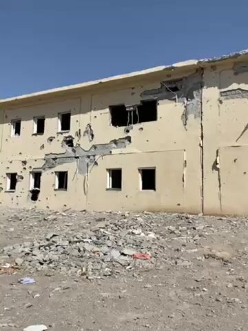The video shows the aftermath of an explosion in what appears to be a war-torn area. A two-story building is visible with extensive bullet holes and structural damage. Debris and rubble cover the ground in front of the building. A white car, severely damaged and burnt, sits amidst the wreckage. There are no people visible in the video. The sky is clear and blue.