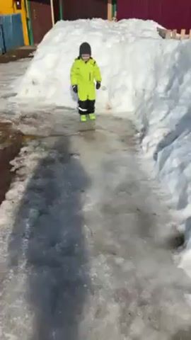 A young child in a bright green snowsuit and black hat is playing with a black and yellow soccer ball on a sunny day in an area with melting snow and ice. The child, who appears to be walking on a path of ice between snowdrifts, kicks the ball and then tries to stop it. Behind the child is a yellow house with white-framed windows and a blue fence. Patches of dirt and standing water are visible on the icy path. The child’s movements are slightly unsteady due to the slippery surface.