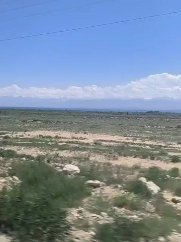This video captures a wide landscape, likely in a rural or semi-arid region of Kyrgyzstan. The dominant subjects are the vast, open terrain under a clear blue sky dotted with white, fluffy clouds. The scene is characterized by dry, sandy patches interspersed with areas of low-lying green vegetation, possibly grasses or scrub. In the distance, a flat, hazy horizon suggests a large body of water or a very flat expanse of land, beyond which, a range of mountains can be faintly seen, their peaks possibly capped with snow. The time of day appears to be daytime, given the bright sunlight and the clear sky. The overall mood is one of spaciousness and natural beauty. There are no people or man-made objects clearly visible in the foreground, though there's a hint of a power line stretching across the top of the frame, suggesting some infrastructure is present in the broader area. The video appears to be filmed from a moving vehicle, creating a sense of travel and progression through the landscape. The activity is simply the observation of this expansive natural environment.