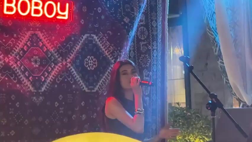 A female singer stands center frame, holding a microphone to her lips. She has long, dark hair and is wearing a black sleeveless top. Behind her is a large, patterned tapestry, possibly a rug, with a red neon sign spelling "BOBOY" in the upper left corner. The lighting is dynamic, with spotlights changing color from red to green to blue. To the right, partially visible, is a microphone stand. Further to the right, a person in a white shirt is also visible. The setting appears to be an indoor or covered outdoor venue, possibly a performance space.