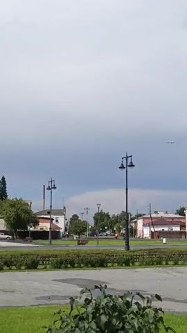 A video shows an airplane flying high in the sky over a town. In the foreground, there is a park area with green grass and bushes, lampposts, and a paved walkway. In the background, there are several buildings, including a large yellow building with white accents and other smaller buildings. A car is parked in front of the yellow building. The sky is partly cloudy.