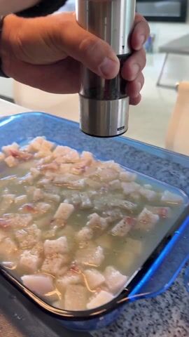 A hand grinds pepper over diced white fish in a blue glass baking dish filled with a light liquid. The pepper grinder is stainless steel with the word "home" printed on it. The dish sits on a countertop with a similar dish beside it. The background shows a modern living room with light-colored furniture. A green cutting board is visible in the foreground.