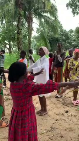 In an outdoor setting with trees, a group of approximately 15 people is engaged in a festive activity, likely a celebration or gathering. The majority of the group appears to be children and young adults, predominantly female, wearing casual clothing and some with headscarves. They are arranged in a loose circle, actively dancing and gesturing with their arms raised. 

Several men are present, playing musical instruments, specifically drums. They are seated and focused on their performance. The setting seems to be a rural or semi-rural area, characterized by a dirt ground with scattered vegetation and simple structures in the background, including a building with a corrugated iron roof.