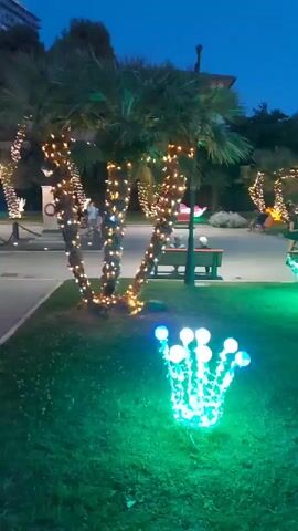 The video shows a park at dusk, decorated with numerous Christmas lights. In the foreground, there is a cluster of grass with a lit green crown-shaped decoration. To the left, a palm tree is wrapped in yellow and white lights, and behind it, another palm tree is also illuminated. In the midground, there are several large, lit decorations resembling glowing blue and green flowers or abstract shapes. Further in the background, people are walking and sitting, and more palm trees decorated with lights are visible, creating a festive atmosphere. The sky is dark blue, indicating it is evening.