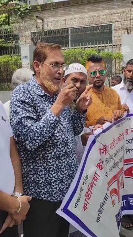 The video shows a group of men standing outside, holding up a large banner. The banner is white with purple and red writing. The text on the banner appears to be in Bengali. The men are dressed in casual clothing. One man in the front is speaking into a microphone. The environment appears to be urban, with a building and fence in the background.