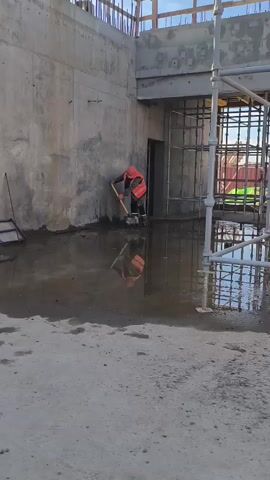 A construction worker wearing an orange hard hat and a reflective orange vest is using a shovel to clear water from a flooded construction site. The worker is standing in a pool of murky water and appears to be shoveling the water towards the left side of the frame. Behind the worker is a concrete wall and scaffolding. The floor of the construction site is concrete, and there are puddles of water scattered around. The lighting suggests it is daytime.