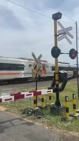 A train passes behind a railroad crossing. The crossing has a red and white horizontal barrier, and a pole with two flashing red lights and a sign that reads "Awas Kereta Api". The train is white with orange stripes. The sky is partly cloudy. The crossing is surrounded by green grass.