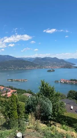 This is a vertical video shot from an elevated vantage point overlooking a large, calm body of water that appears to be a lake. Several islands dot the lake, with buildings visible on some of them, including a prominent structure with a red roof on one island. The water is blue and reflects the sky, with some boats visible on the surface. The distant shoreline is lined with towns and dotted with houses and vegetation, and is backed by rolling, tree-covered hills that transition into larger mountains in the background. The sky is a clear, bright blue with scattered white clouds. In the foreground, lush green foliage and trees frame the view, with some houses and a section of a roof with solar panels visible on the right side. The overall impression is a scenic, sunny day at a lake resort or coastal area.