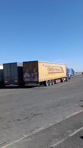 The video shows a parking area with several large trucks and a white van. The trucks are parked in a line, and the van has its doors open. The sky is clear and blue, and the sun is shining. The ground is paved with asphalt. In the background, there is a road and some vegetation.