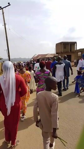 The video shows a large group of people walking down a street in a residential area. The street is unpaved and bordered by buildings and a power pole. Most of the people are dressed in brightly colored clothing and many of the women are wearing head coverings. The camera is positioned behind the group of people, providing a view of their backs. The sky is overcast and the overall atmosphere is peaceful.
