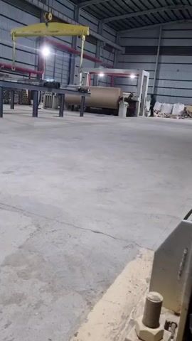 The video shows the interior of a large industrial building. A large roll of brown material sits on a machine in the center. A yellow crane hangs overhead. A person stands near the machine. There are rows of pipes running along the ceiling and stacks of materials in the background. The floor is concrete with cracks and some dirt. In the foreground, part of another machine is visible. The scene is well-lit.