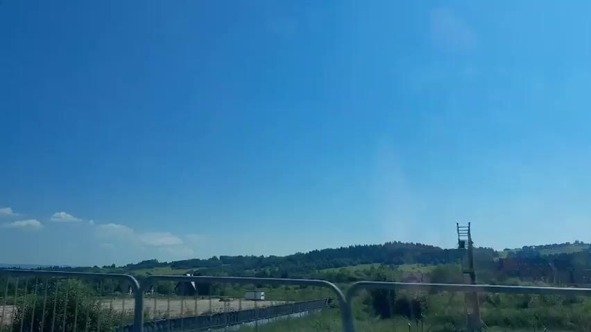 This media file captures a scenic view of a rural landscape under a bright blue sky. The foreground features a metal railing, suggesting the vantage point is from a bridge, walkway, or elevated structure. Beyond the railing, vegetation and some structures are visible, indicating a settled area within a natural setting. The midground is dominated by rolling hills covered in dense green forests, characteristic of a mountainous or hilly region. The sky is a clear, vibrant blue with only a few wispy white clouds scattered across it, suggesting fair weather and a sunny day. The time of day appears to be midday or early afternoon due to the bright sunlight and clear sky. The overall impression is one of tranquility and natural beauty, with the vast expanse of the sky and the lush greenery of the hills creating a peaceful and expansive atmosphere. The location context of Bańska Niżna, Poland, places this scene in a European rural environment, likely in a mountainous or sub-mountainous area known for its natural landscapes. There is no visible text within the media file.