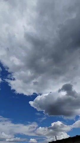 The video shows a sky with dynamic cloud formations. The clouds range from bright white and fluffy to dark gray, indicating potential changes in weather. A portion of a blue sky is visible between the clouds. In the bottom right corner of the frame, the edge of a dark-colored structure, possibly a roof or building, is visible. There are no people or animals present in the video. The camera angle appears to be looking upwards.